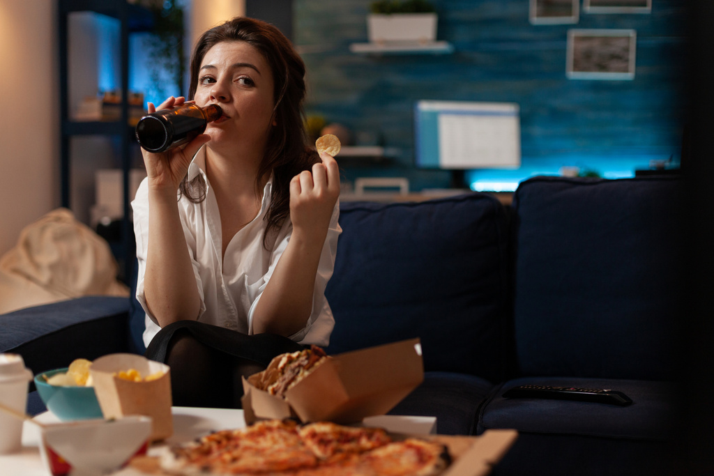 woman-dressed-casual-drinking-beer-from-bottle-while-holding-potato-chip-looking-television-comedy-sitcom-show-person-sitting-co